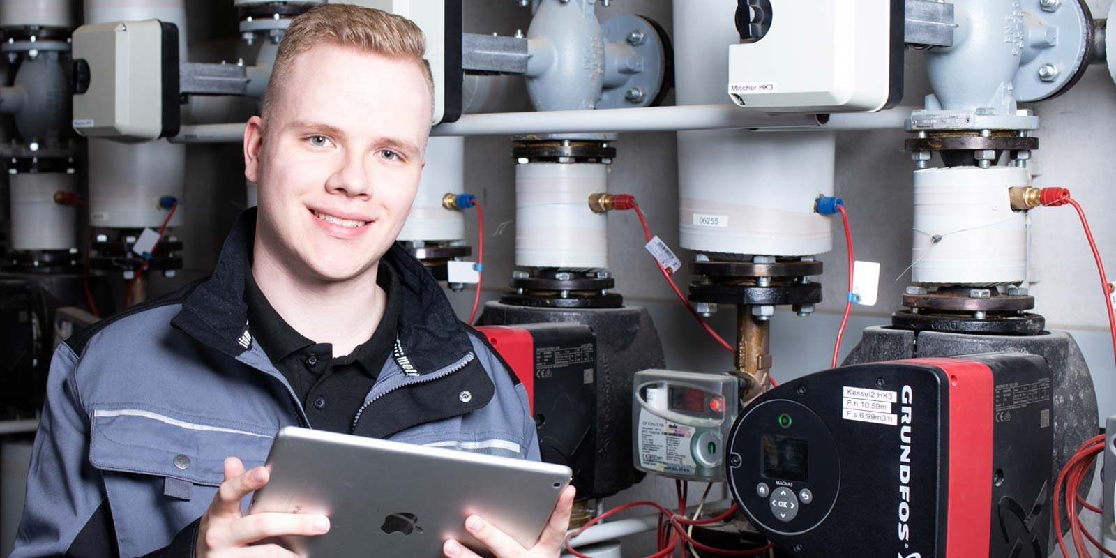 Ein junger Anlagenmechaniker vor einer Heizungsanlage, der ein Tablet hält